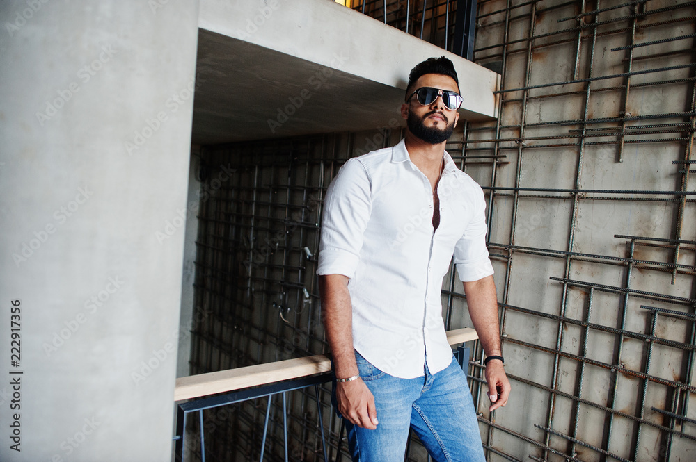 Stylish tall arabian man model in white shirt, jeans and sunglasses posed against steel wall indoor. Beard attractive arab guy.