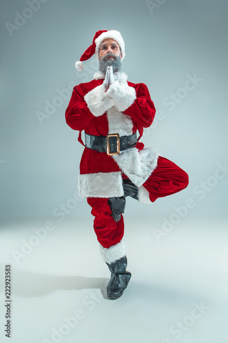 Funny serious guy with christmas hat sitting at yoga pose at studio. New Year Holiday. Christmas, x-mas, winter, gifts concept. Man wearing Santa Claus costume on gray. Copy space. Winter sales.