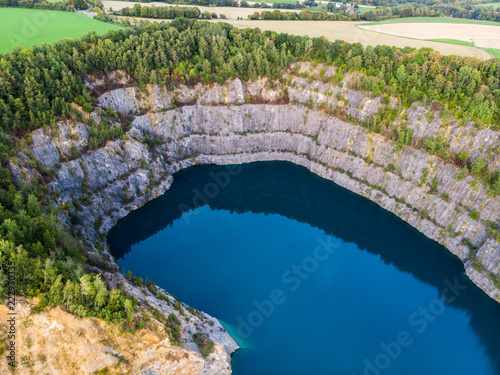 (Laufaufnahme, Drohne) Felswand eines Steinbruchs mit blauem See im Vordergrund