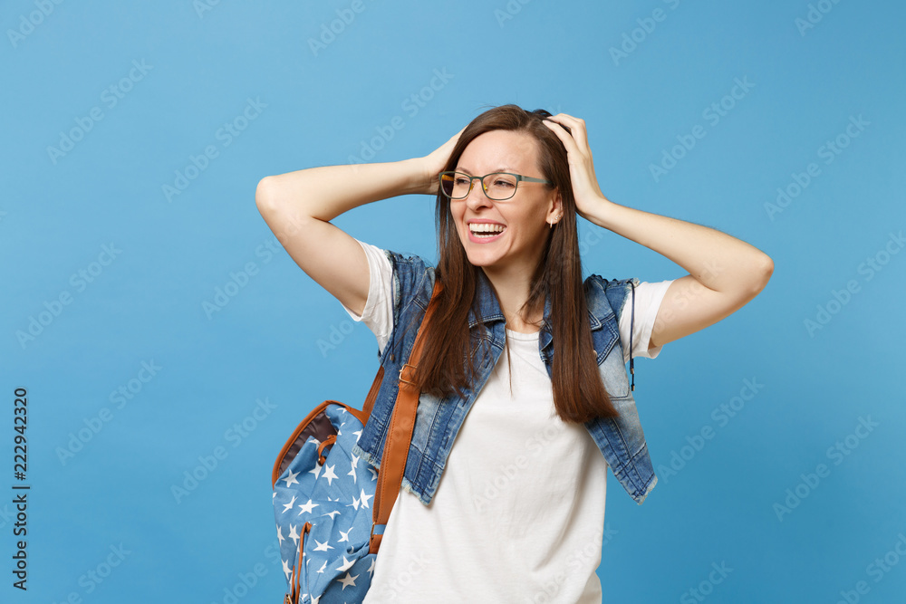 Portrait of young happy laughing woman student in denim clothes glasses with backpack look aside keeping hands on head isolated on blue background. Education in high school university college concept. Portrait of young happy laughing woman student in denim clothes glasses with backpack look aside keeping hands on head isolated on blue background. Education in high school university college concept.