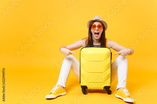 Traveler tourist woman in summer casual clothes, hat sit with suitcase isolated on yellow orange background. Female passenger traveling abroad to travel on weekends getaway. Air flight journey concept