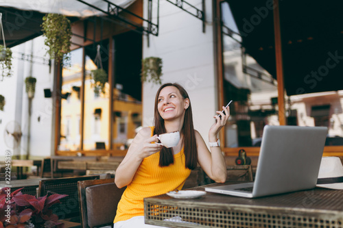 Happy girl in outdoors street coffee shop cafe sitting at table with laptop pc computer, texting message on mobile phone, drink cup tea in restaurant during free time. Mobile office freelance concept. © ViDi Studio