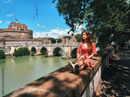 Brunette girl in red dress, striped sits on the embankment of a Roman bridge across the Tiber river, overlooking the huge Roman monuments architecuture photo