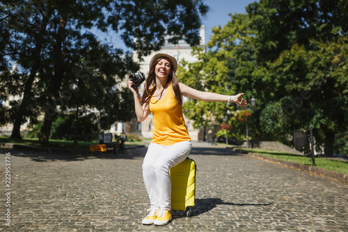 Overjoyed traveler tourist woman in hat sitting on suitcase holding retro vintage photo camera spreading hands in city outdoor. Girl traveling abroad on weekend getaway. Tourism journey lifestyle.