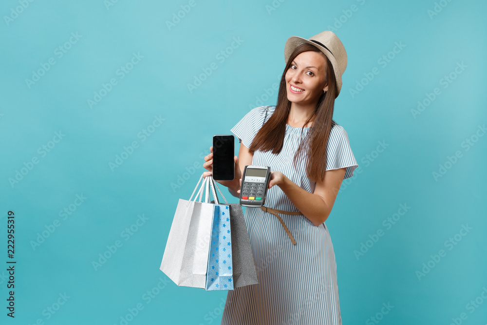 Portrait woman in dress, hat holding packages bags with purchases after shopping, wireless modern bank payment terminal to process and acquire credit card payments isolated on blue pastel background. Portrait woman in dress, hat holding packages bags with purchases after shopping, wireless modern bank payment terminal to process and acquire credit card payments isolated on blue pastel background.