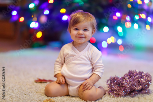 Adorable baby girl holding colorful lights garland in cute hands. Little child in festive clothes decorating Christmas tree