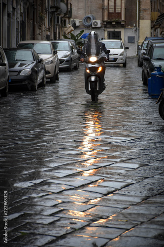 Man riding a small motorbike through the streets of an italian city, with the headlinght reflecting in the wet stone pavement of the street