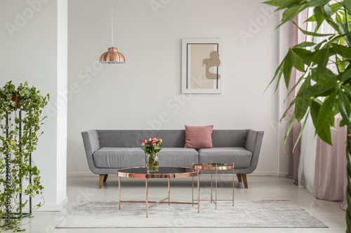 Close-up of leaves in an elegant living room interior with a grey couch and copper tables decorated with roses. Real photo