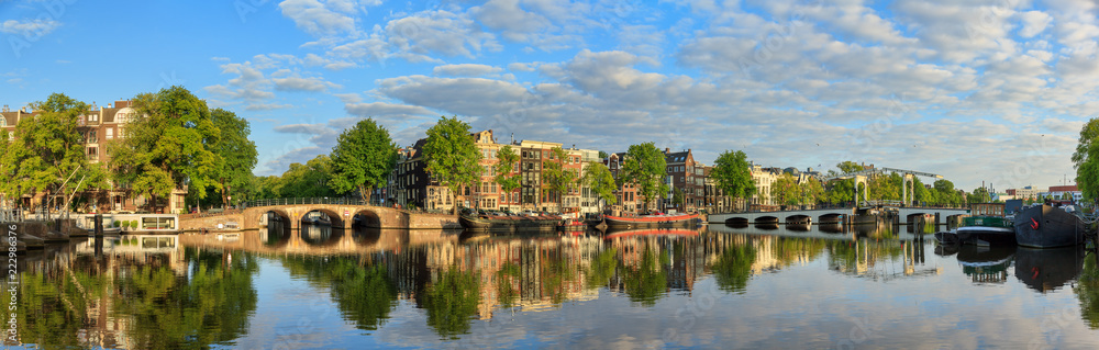 Fototapeta premium Piękna panoramiczna panorama rzeki Amstel i chudy most (Magere brug) w Amsterdamie, Holandia, w słoneczny letni poranek z chmurami i lustrzanym odbiciem