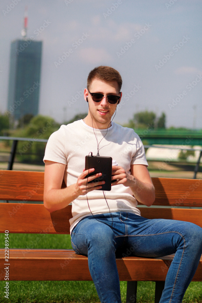 Obraz premium Handsome boy wearing sunglasses using tablet and headphones, sitting on a bench and drinking coffee to go. Handsome student immersed in social media. Background scenery bridge, river and green grass.