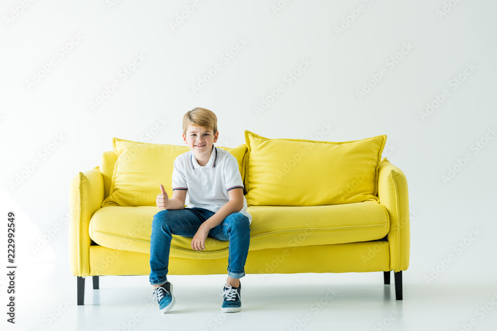 smiling adorable boy sitting on yellow sofa and showing thumb up on white