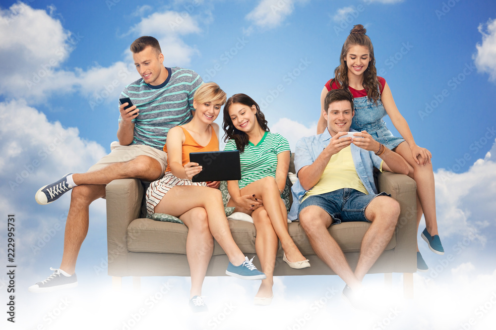 Obraz premium friendship, leisure and technology concept - group of happy smiling friends with tablet pc computer and smartphones sitting on sofa over blue sky and clouds background