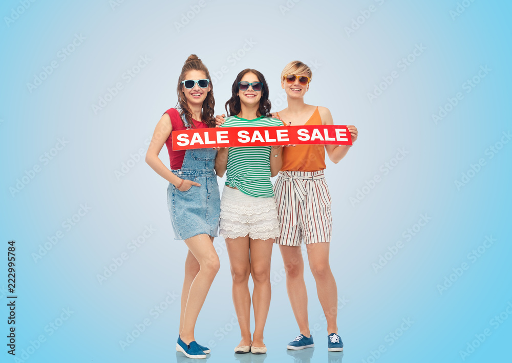 Fototapeta premium shopping and people concept - happy smiling teenage girls with sale banner over blue background