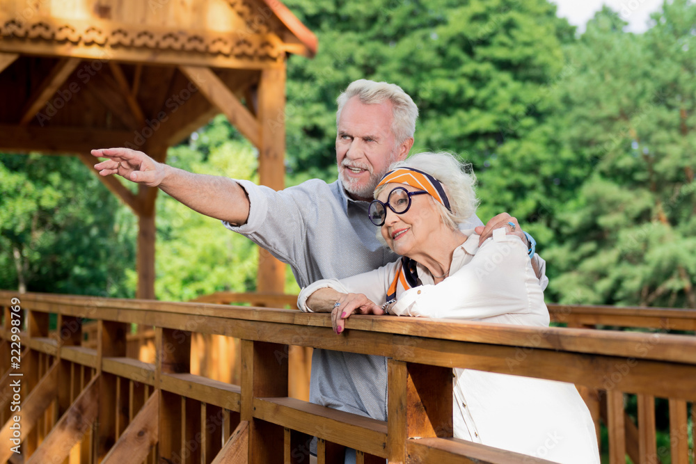 Showing direction. Calm confident senior man standing on the bridge with his smiling wife and pointing into the distance Showing direction. Calm confident senior man standing on the bridge with his smiling wife and pointing into the distance