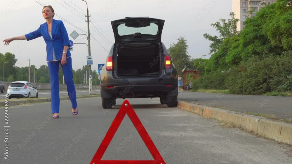 Woman in suit stands near black car and holds her hand up. She seeks about help. Her car is broken. Girl is upset. Woman wants to stop somebody in car to help her. Woman in suit stands near black car and holds her hand up. She seeks about help. Her car is broken. Girl is upset. Woman wants to stop somebody in car to help her.