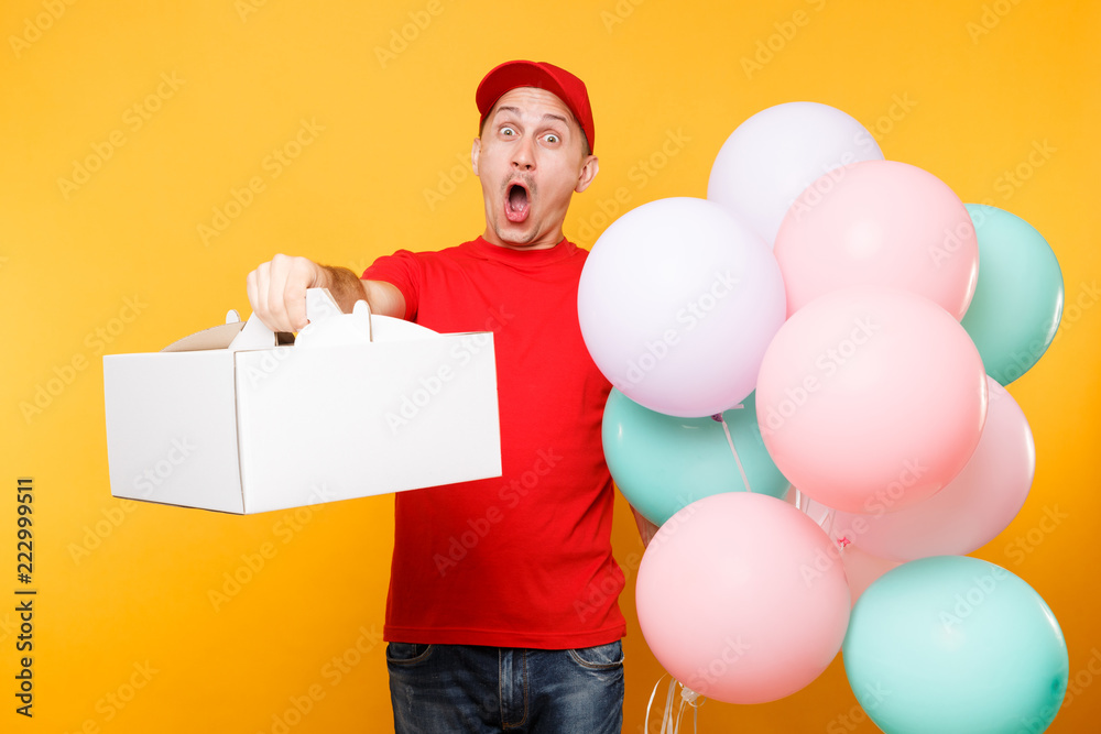 Man giving food order cake box isolated on yellow background. Male employee courier in red cap t-shirt hold colorful air balloons, dessert in empty cardboard box. Delivery service concept. Copy space.