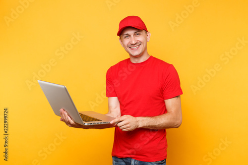 Delivery man isolated on yellow orange background. Professional male employee courier in red cap, t-shirt holding working typing on laptop pc computer. Service concept. Copy space for advertisement.