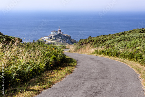 Spain, Finisterre: Famous lighthouse on rocky cliff with road, seascape, ocean sea, skyline and blue sky in the background. It is located on a rock-bound peninsula and was known as end of the world. photo