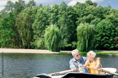 Enjoying boating. Peaceful beautiful senior couple smiling and enjoying boating while hugging in the boat