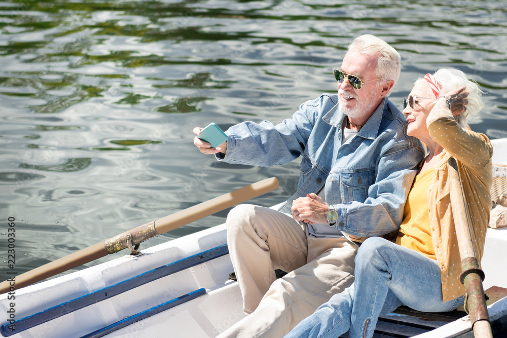 Taking selfies. Positive modern couple of pensioners smiling and taking selfies while sitting in the boat