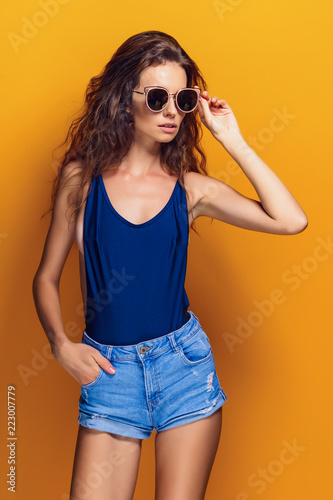 Young sexy woman dressed in swimsuit and blue jeans shorts, sunglasses posing on the yellow background.