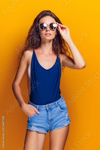 Young sexy woman dressed in swimsuit and blue jeans shorts, sunglasses posing on the yellow background.