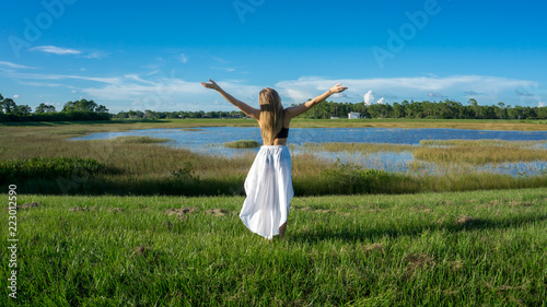 Blonde young woman standing backwards in a beautiful field landscape outdoors with raise hands arms to the sky