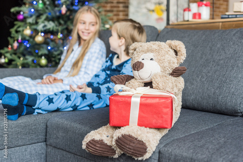 close-up view of teddy bear with christmas gift and kids in pajamas sitting on sofa behind