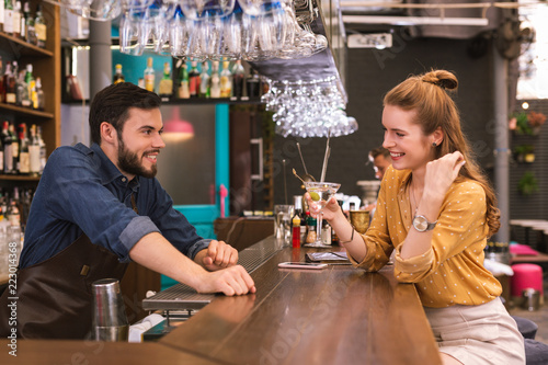 Friendly bar. Positive young girl sitting in the bar and smiling while having positive talk with handsome barman