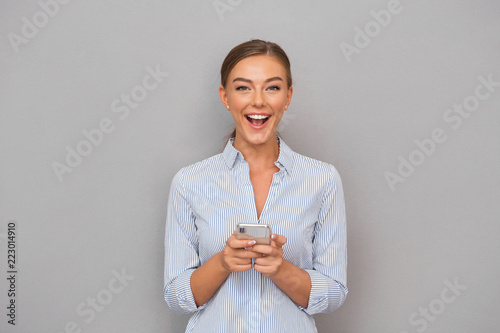 Smiling business woman standing over grey wall background using mobile phone.