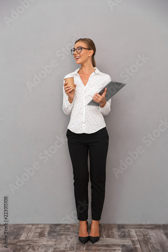 Beautiful business woman standing over grey wall background holding clipboard drinking coffee.