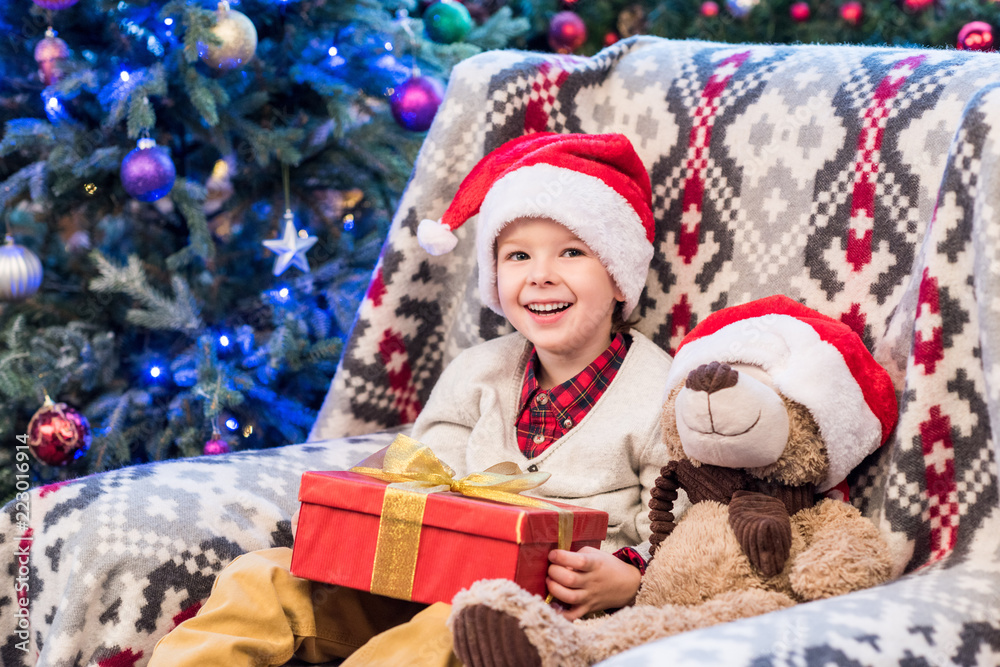 happy little boy in santa hat holding christmas present and smiling at camera