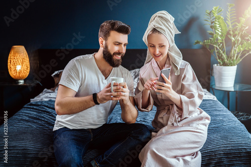 Morning.Young couple,woman after shower in bathrobe and towel on her head, sitting in bedroom on bed. Girl shows information to man on smartphone, guy looks at screen, drinks coffee. Social networks.