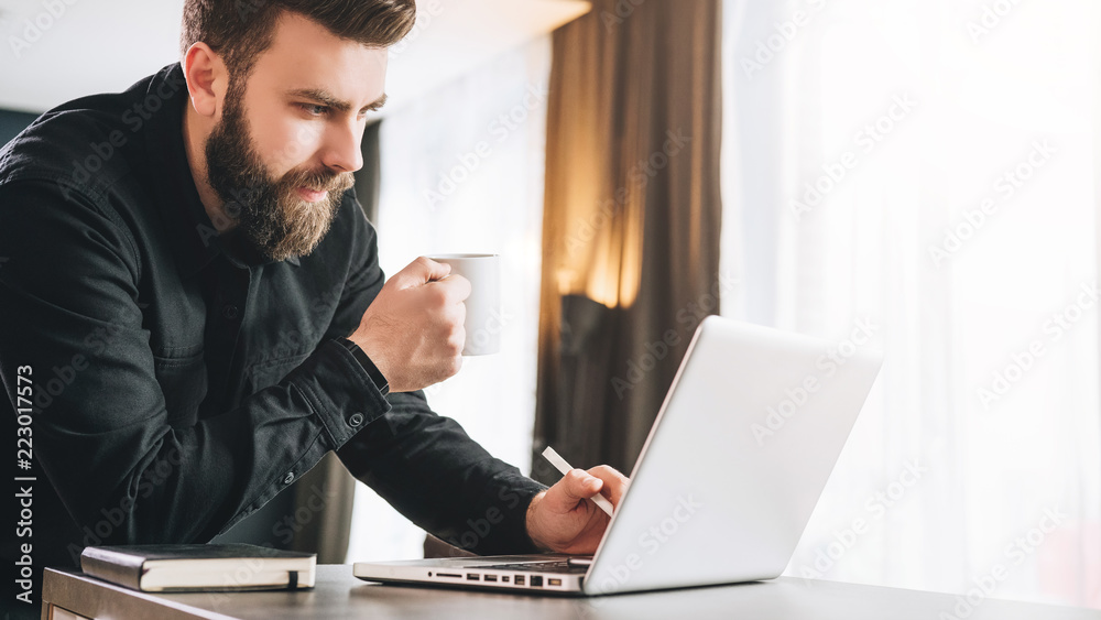 Bearded smiling businessman is standing near table, is drinking coffee, using laptop. Man works in office on computer. Bearded smiling businessman is standing near table, is drinking coffee, using laptop. Man works in office on computer.