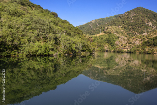 Reflection of a beautiful rural green tree covered hill in calm still lake sea water with riverside bank, mountain chain and blue sky - concept untouched nature environment forest scenic panorama