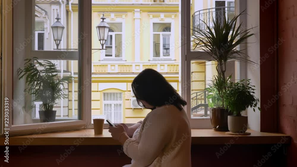 Stout caucasian women scrolling on mobile phone near window in cafe with paper cup Stout caucasian women scrolling on mobile phone near window in cafe with paper cup
