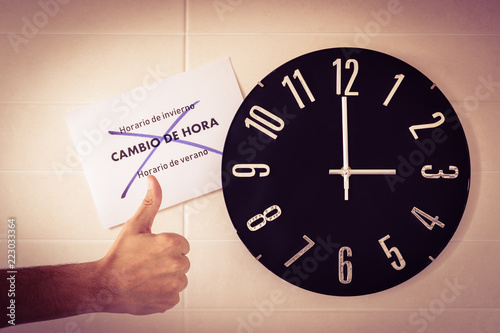 Big black clock on white wall. Time change. DST. Survey of the European Union. Gesture of disagreement . Thumb down of caucasian man. Spanish banner. Translation: winter time, time change, summer time