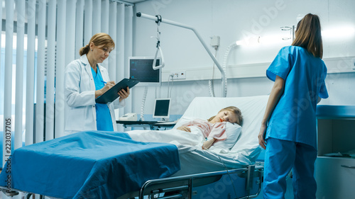Sick Little Girl Lies on a Bed In the Hospital  Friendly Doctor Writes Medical Record  Data into Clipboard  Talks with Nurse. Cute ill Child is Taken Care of in the Modern Pediatric  Children Ward.
