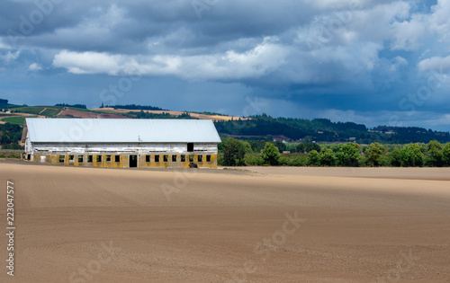 An old cement block shed with a white roof stands out against plowed and groomed fields, hills behind and a cloudy sky above.