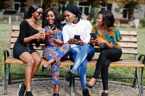 Group of four african american girls sitting on bench outdoor with mobile phones at hands. photo