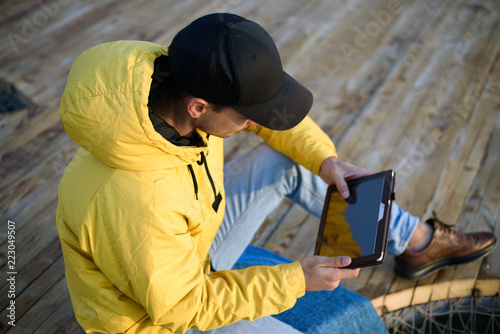Close-up of a man on top in a yellow jacket and a black cap sitting on the pier with a tablet