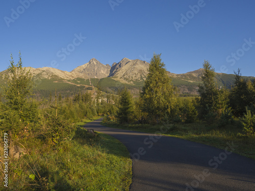 Tatra Mountains range with asphalt road curve, golden hour light, way from Tatranska Lomnice to Start with green trees and grass. Early autumn panorama photo