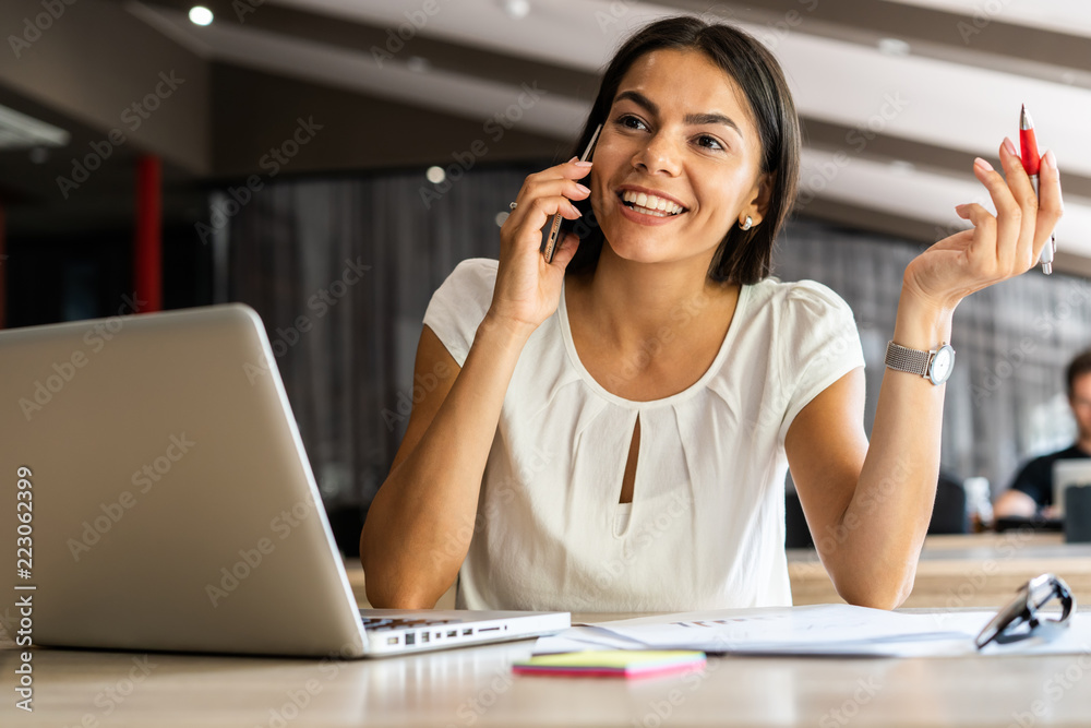 Good business talk. Cheerful young beautiful woman talking on mobile phone and using laptop with smile while sitting at her working place. Good business talk. Cheerful young beautiful woman talking on mobile phone and using laptop with smile while sitting at her working place.