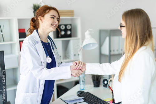 A beautiful young girl in a white coat is standing near the desk in the office and shaking hands with the patient. The patient thanks the doctor.