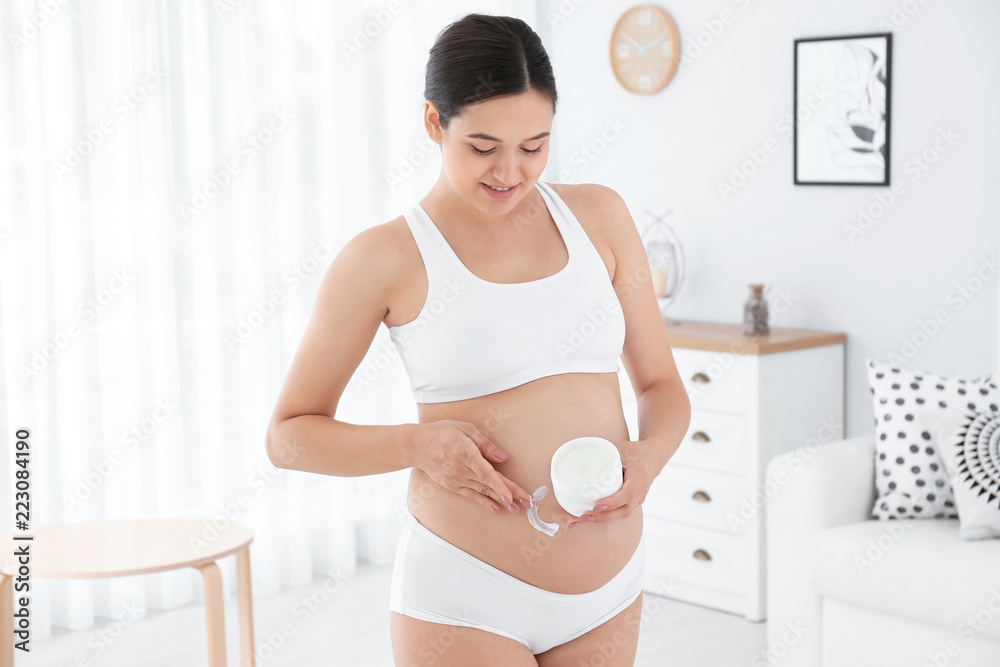 Pregnant woman applying body cream on belly at home