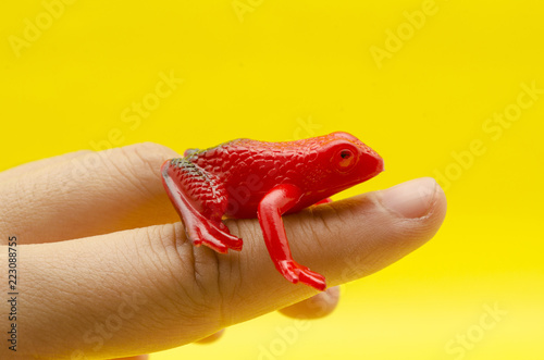 Plastic toy frog on a finger against yellow background. Toy red frog on yellow background