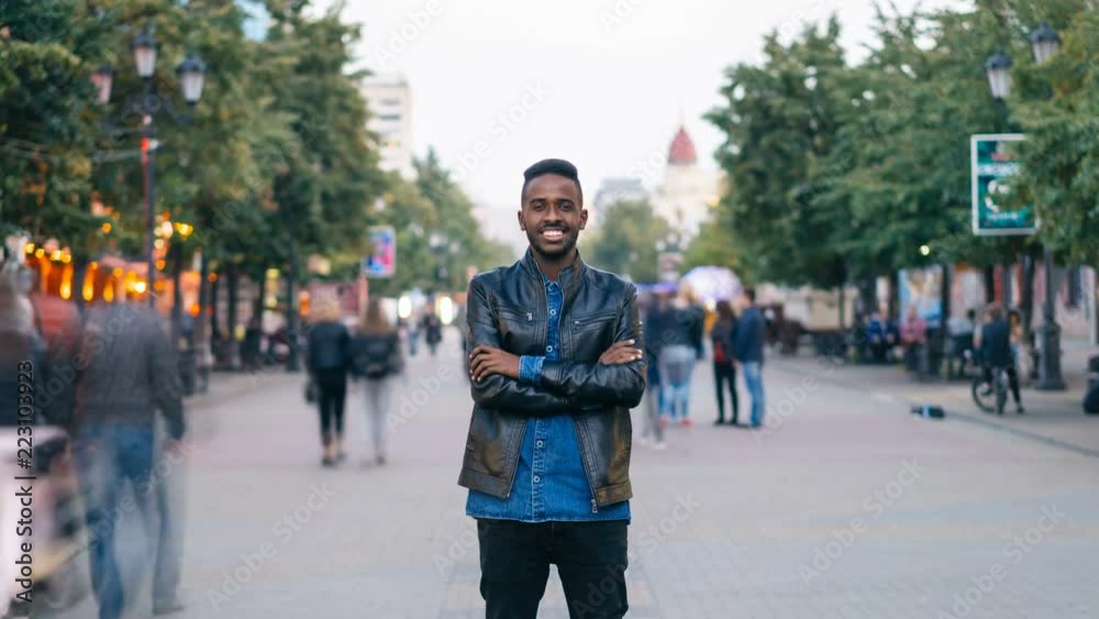 Zoom in time-lapse of happy African American guy wearing jeans and leather jacket standing alone in street downtown, smiling and looking at camera with crowd moving by. Zoom in time-lapse of happy African American guy wearing jeans and leather jacket standing alone in street downtown, smiling and looking at camera with crowd moving by.