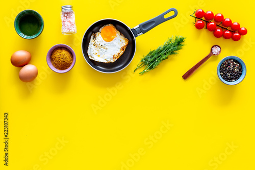 Recipe of fried eggs with vegetables. Ready eggs in a frying pan near cherry tomatoes, greenery, spices, raw eggs on yellow background top view copy space