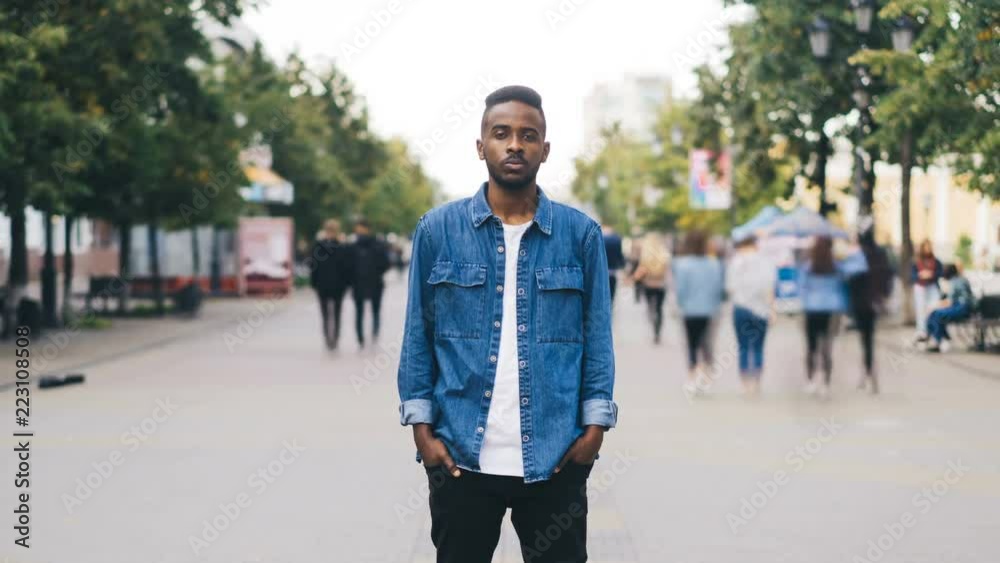 Zoom in time-lapse of unhappy single African American guy standing alone in the city center with hands in hockets and looking at camera with straight face.