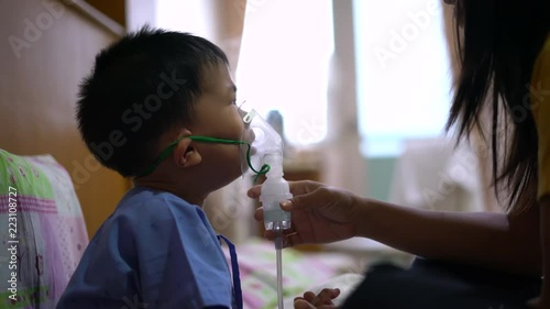 Sick Asian boy about 2 years and 8 months in hospital uniform using inhaler containing medicine to stop coughing from disease like flu or RSV, Respiratory Syncytial Virus